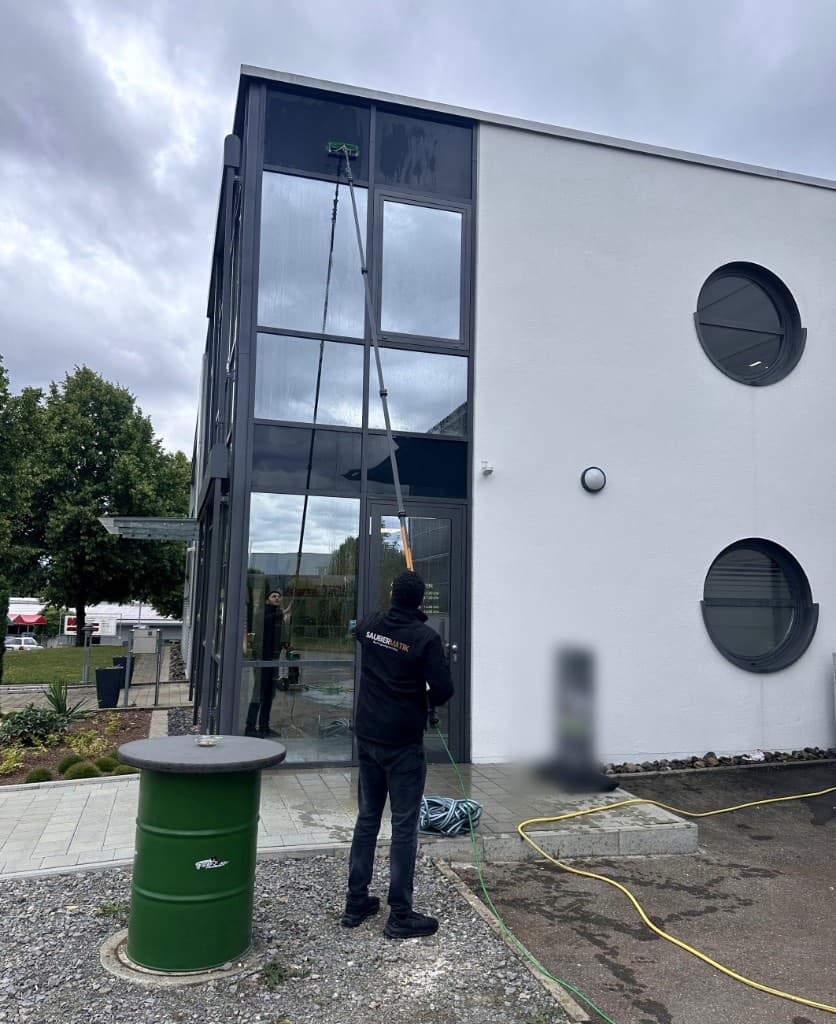 Saubermatik-Mitarbeiter reinigt Fenster mit Teleskopstange und Bürste an einer Glasfassade
